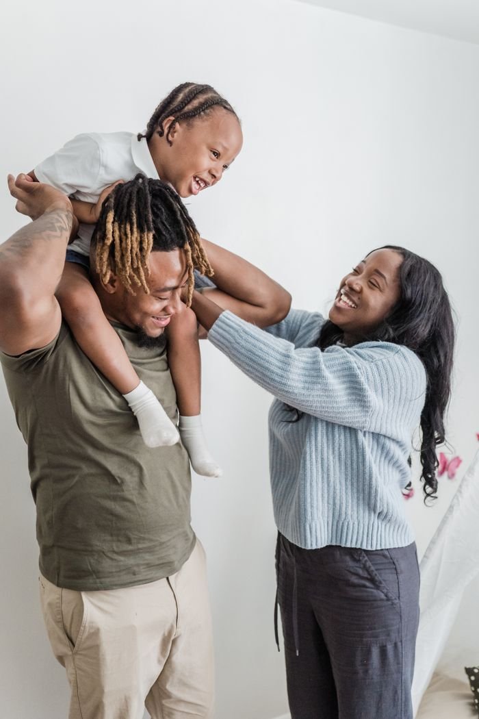 Joyful family moment with parents lifting child onto shoulders, laughing indoors.