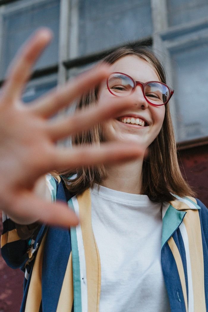 Happy teenage girl with glasses smiling and waving outdoors in a playful moment.